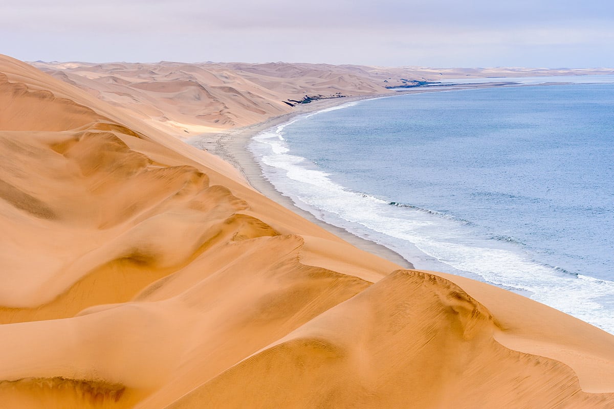 A vivid landscape where towering golden sand dunes meet the cool blue ocean of Namibia's Skeleton Coast. The dunes create sweeping patterns, leading to the shoreline which is marked by gentle waves, showcasing