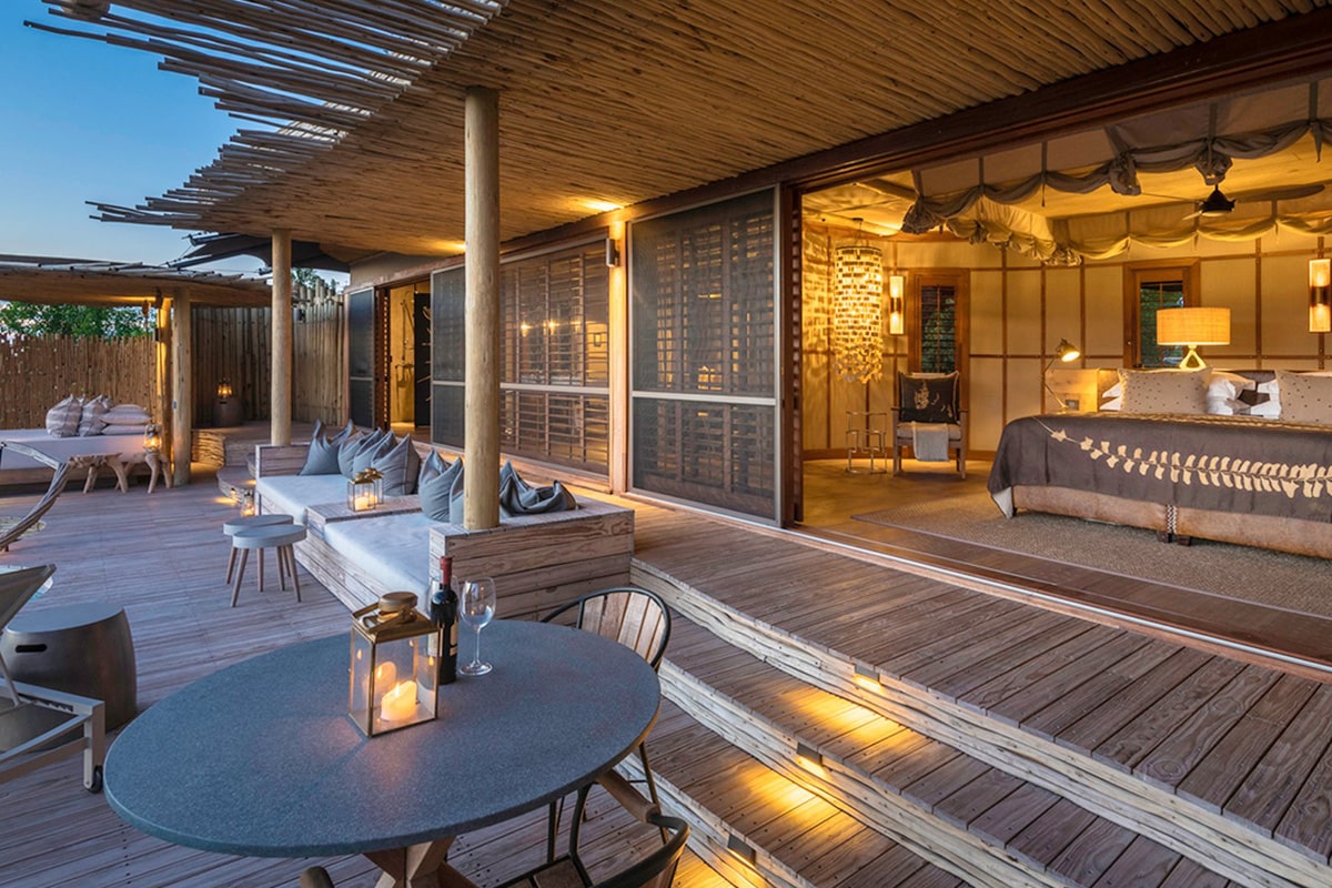 Luxury outdoor patio and bedroom of a luxury safari lodge in Botswana at dusk, featuring elegant wooden flooring, comfortable seating with plush cushions, a bed with delicate netting, and soft atmospheric lighting.