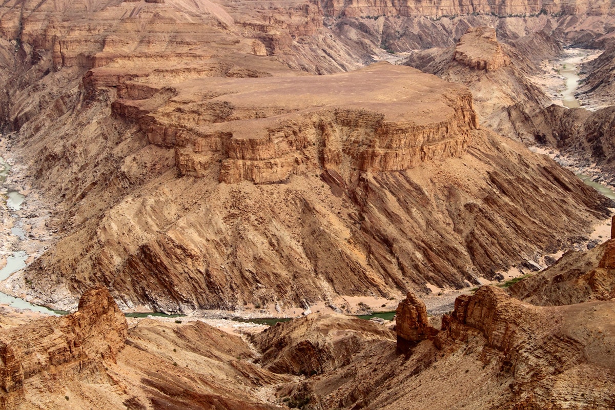 Aerial view of a rugged canyon with layered sedimentary rock formations in various shades of brown and tan. A winding river cuts through the bottom of the canyon, surrounded by sparse green vegetation. Discover this