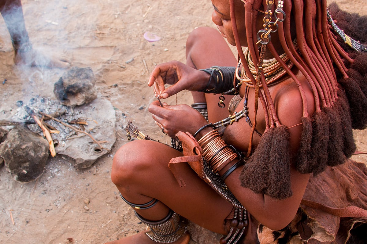 A Himba woman from Namibia, adorned in traditional orange ochre body paint and jewelry, sits by a small fire, crafting an item with her hands. Beaded ornaments and metal bracelets encircle