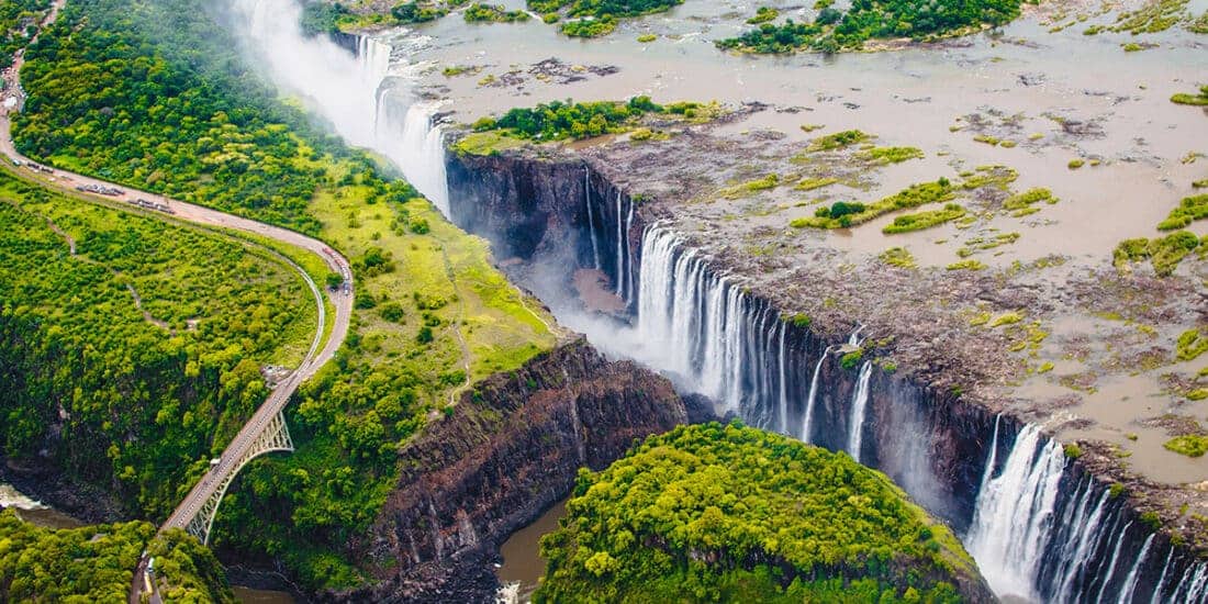 Aerial view of Victoria Falls in Zambia with water cascading down cliffs surrounded by lush vegetation. A train runs on a track beside the river, and a bridge spans the gorge near mist clouds. Nearby