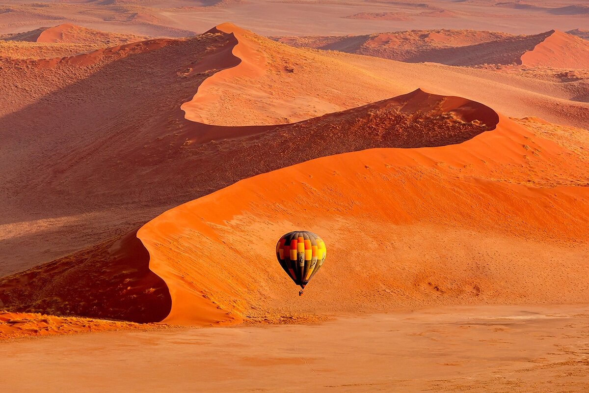 A lone hot air balloon with a patchwork design of black, yellow, and orange floats above the vast orange sand dunes of a desert. The sun illuminates the dunes, accentuating their ridges and casting shadows that highlight their curves and textures—an incredible experience in travel Namibia.