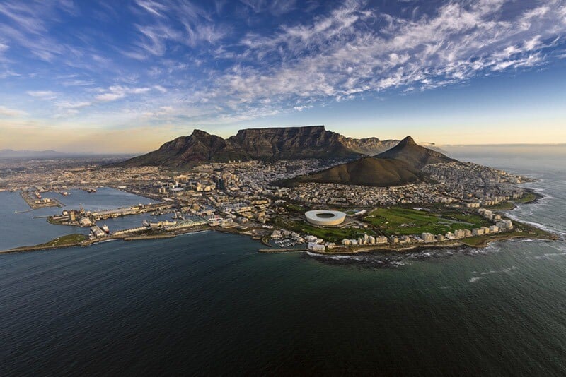Aerial view of Cape Town, South Africa showing Table Mountain with its flat summit and the cityscape below, including the distinctive circular stadium near the coastline, under a clear sky.