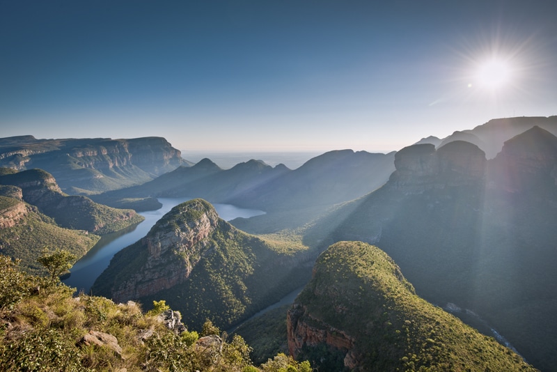 Sunrise over a serene river winding through the mountainous landscape of the Panorama Route, with rugged cliffs and lush greenery under a clear blue sky illuminated by a breathtaking sun.