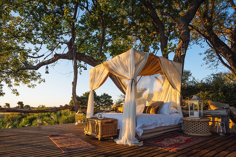 Outdoor safari-style bedroom setup on a wooden platform with a canopy bed draped in sheer white curtains, surrounded by lush trees during sunset, decorated with soft pillows, a rug, and a small lantern at a