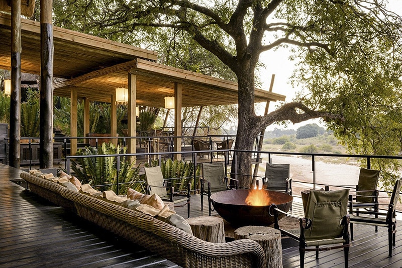 A serene outdoor deck of a luxury safari lodge with a fire pit in the center, surrounded by rattan chairs and cushions, overlooking a lush, tree-lined landscape at dusk in Kruger National Park.