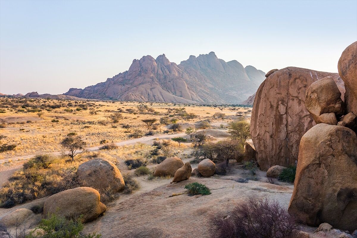 A desert landscape at sunrise featuring a range of rugged mountains in the background and large rounded boulders in the foreground. Sparse vegetation is scattered across the sandy plains of Namibia.