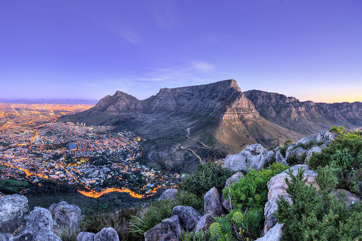 Panoramic view of Cape Town, South Africa, at dusk from a high vantage point, showcasing the illuminated cityscape to the left and the majestic Table Mountain under a clear sky to the right