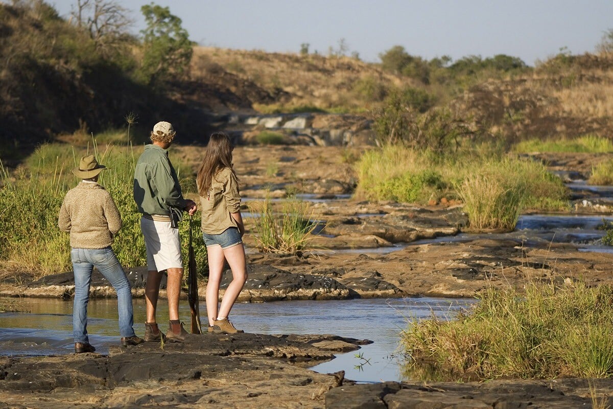 Three people standing near a river in the Hoedspruit area of the Kruger region, observing the surroundings. Two men and one woman are wearing safari outfits, surrounded by rocky terrain and sparse vegetation