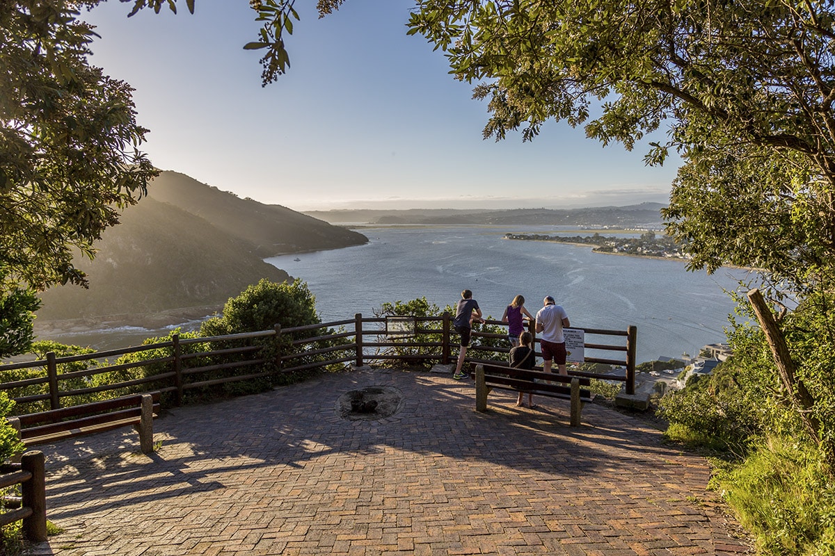 Three people stand at a lookout with a wooden railing overlooking a scenic river estuary. The sunset casts a warm glow over the water, surrounded by lush hills. The foreground features a paved area with trees