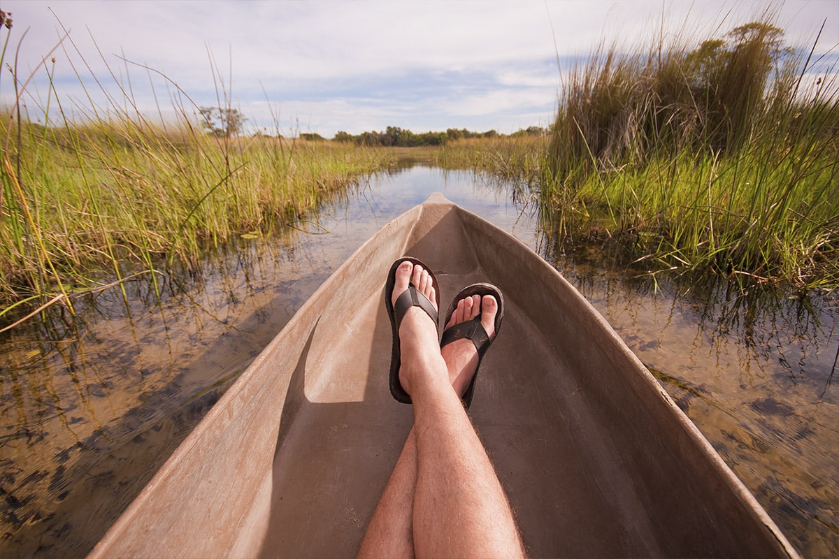 A person's view from a canoe showing their legs and feet in sandals, as they paddle through a grass-lined waterway in Southern Africa under a clear sky. The perspective emphasizes the tranquility and narrow
