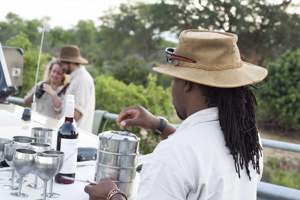 A man in a safari hat with sunglasses observes a couple taking a photo with a smartphone by a vehicle on an African Safari, with wine and metal cups on a table nearby.
