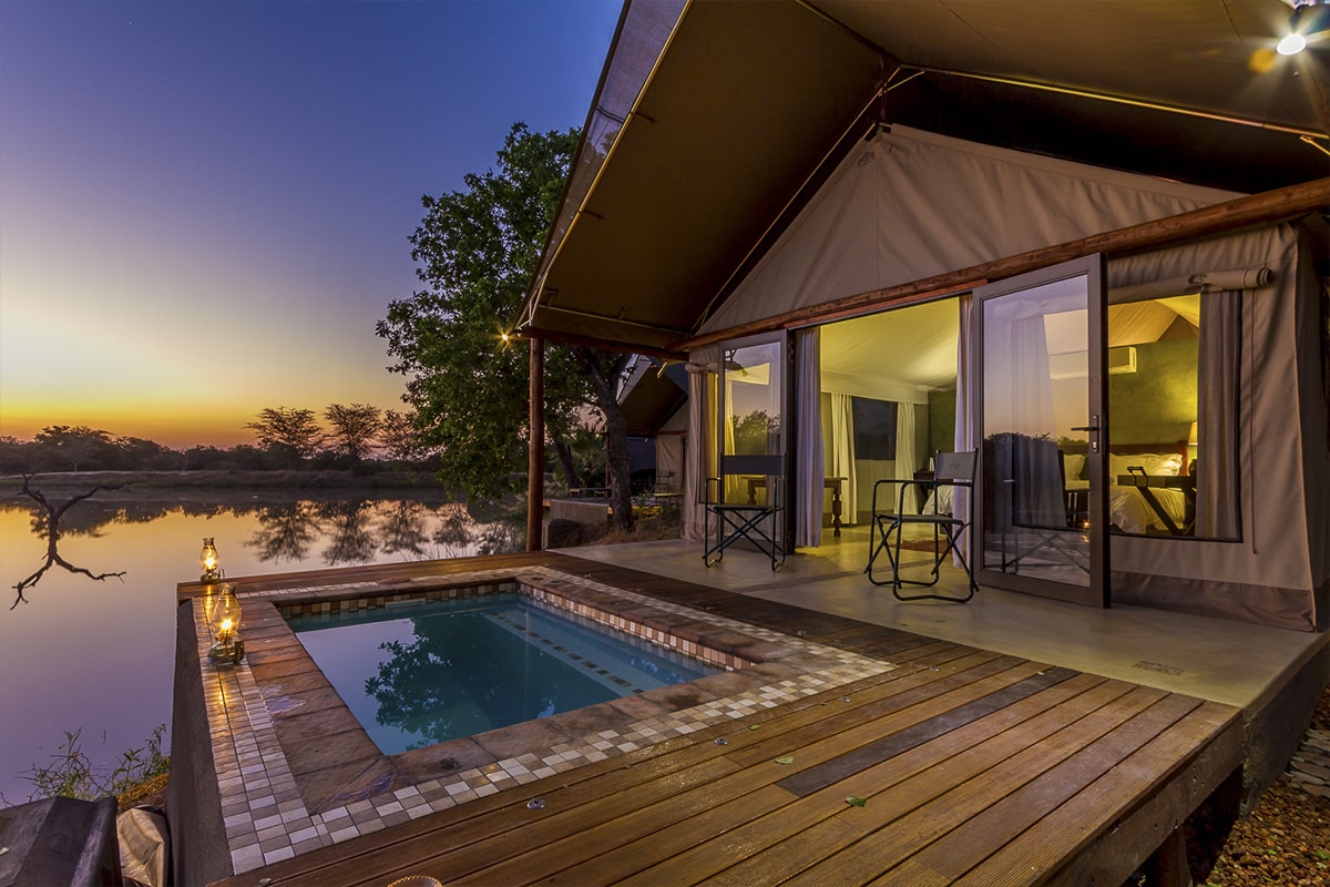 Luxurious African Safari lodge tent with a private pool on a wooden deck overlooking a tranquil river at sunset, featuring vibrant sky colors and serene natural surroundings.