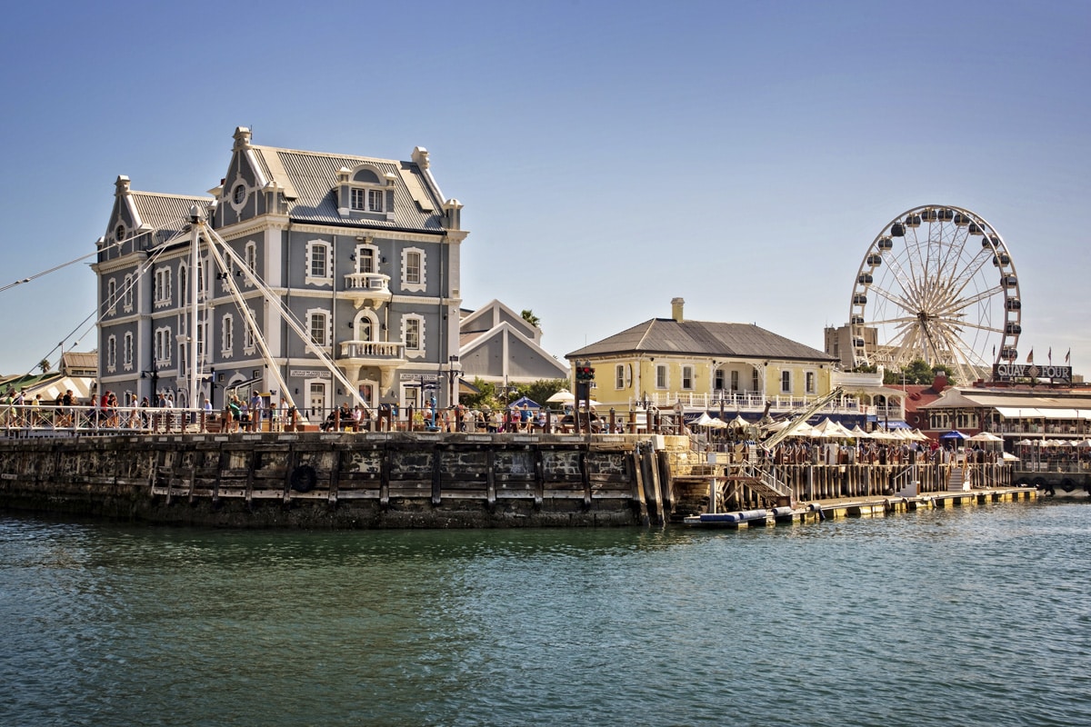 Image of a vibrant waterfront scene featuring a large, stately grey building with white trim on the left. To the right, there's a yellow building with a ferris wheel in the background. A crowd is visible near the buildings, enjoying the sunny day along this popular travel destination in South Africa.