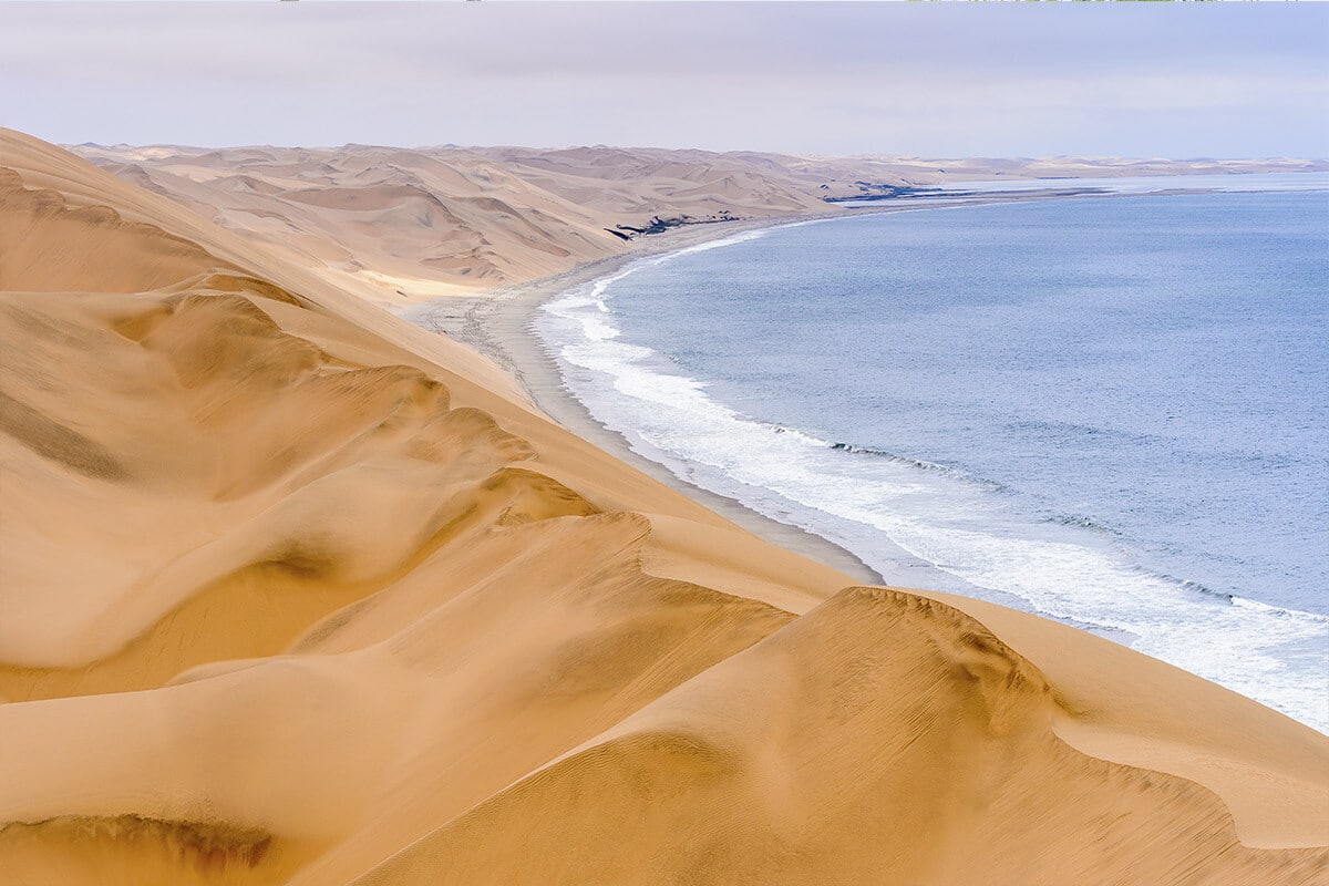 A panoramic view of the Namib Desert coastline near Swakopmund showcasing a series of large golden sand dunes cascading down to a tranquil blue ocean. Waves gently lap against the shoreline, contrasting