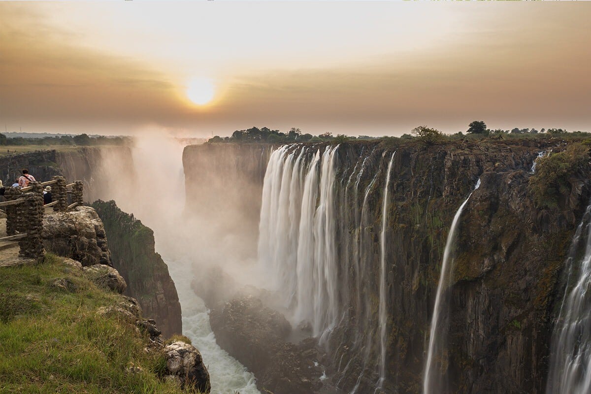 Sunset over the majestic Victoria Falls with dense mist rising from the bottom. A couple sits on a cliff edge, viewing the waterfall that spans the horizon, marked by rugged cliffs and lush vegetation.
