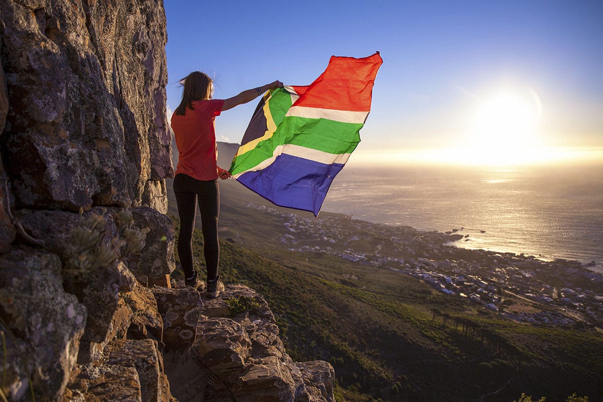 A person stands on a rocky ledge overlooking a vast seascape at sunset, holding up a large South African flag that flutters in the breeze, embodying the spirit of travel in South Africa.