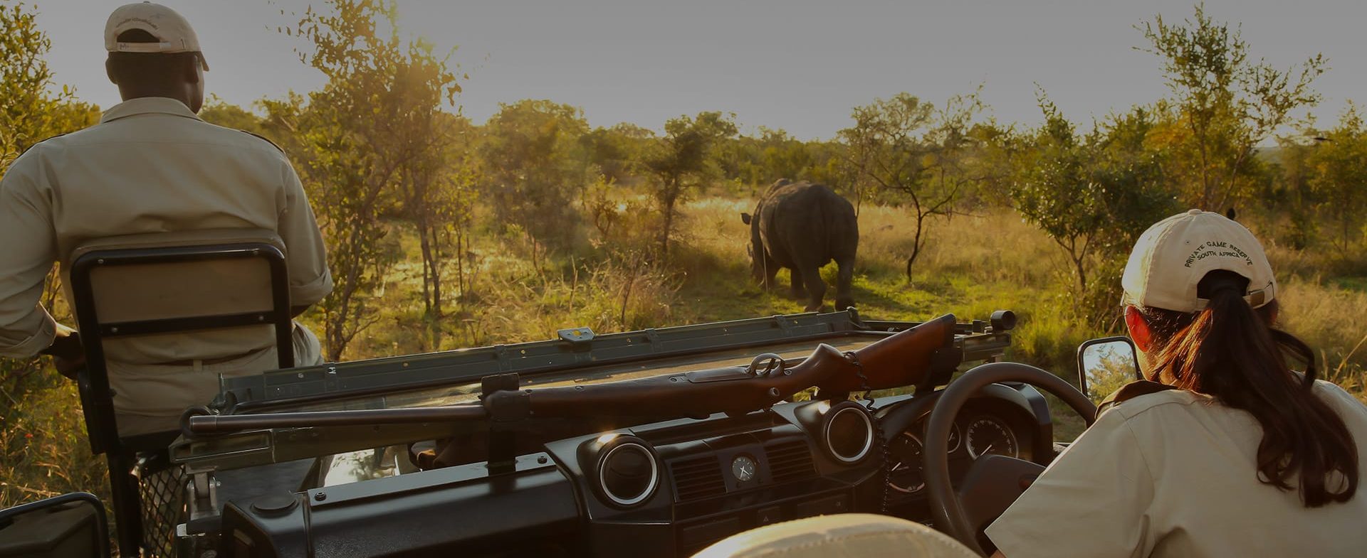 Two people, one wearing a hat and the other a cap, are seated in a luxury safari vehicle watching a distant rhinoceros in a grassy savannah during sunset.