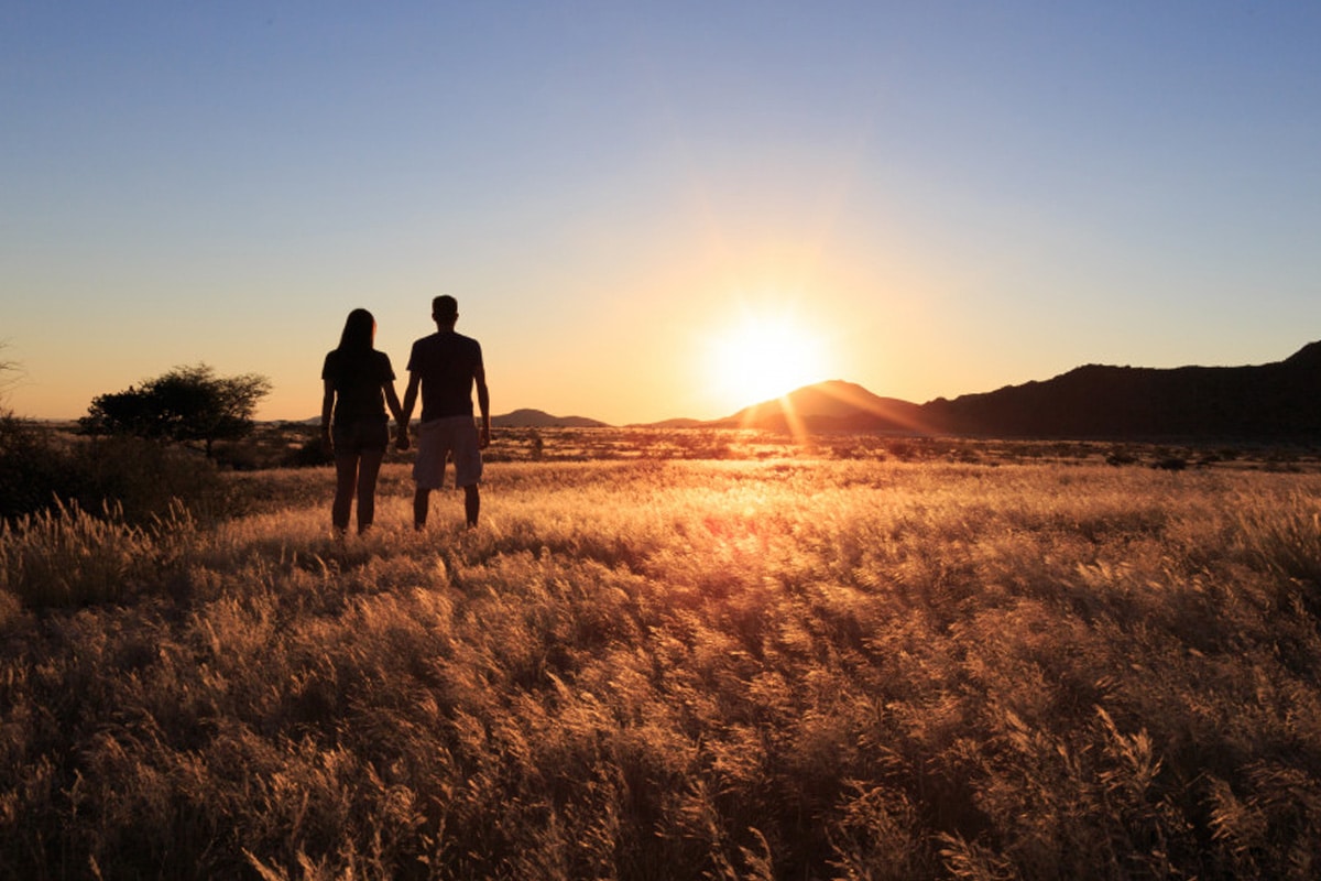 A couple holding hands in a field at sunset, silhouetted against a bright sky. The setting sun casts a warm glow over the landscape, highlighting the textures of the grass and distant hills,