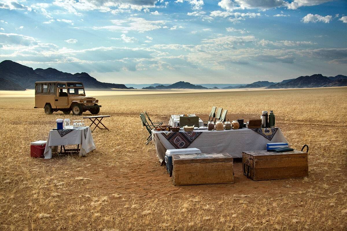 A meticulously arranged outdoor dining setup in a vast desert, featuring a table set with dishes and a vintage 4x4 vehicle parked nearby, surrounded by distant mountains under a vast sky during your Luxury Nam