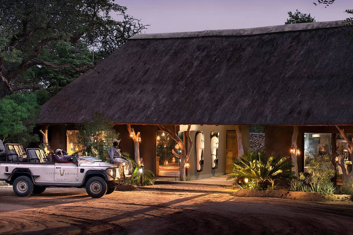 Two people in a safari vehicle parked outside a luxury lodge illuminated by warm lights at dusk, with lush plants surrounding the entrance.