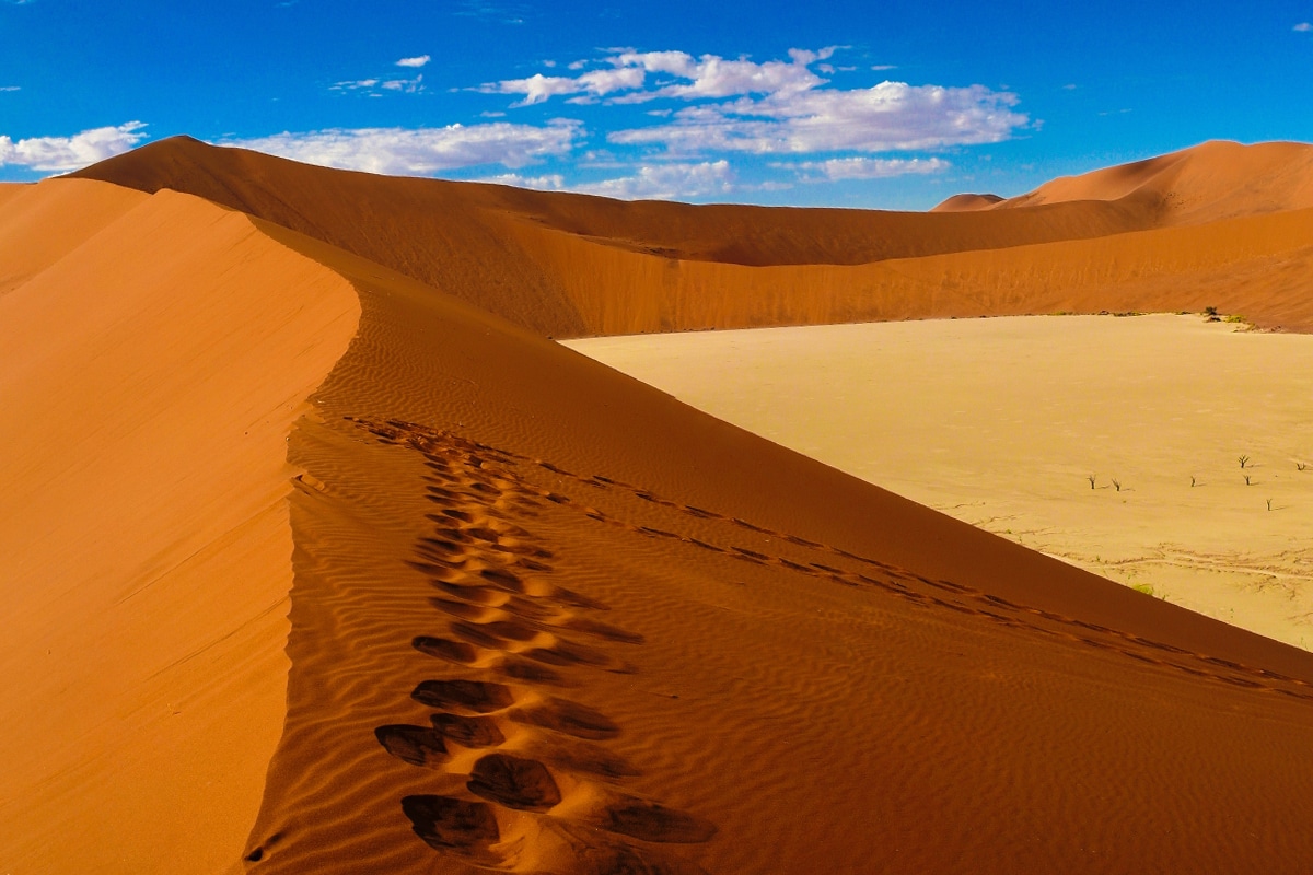 Vivid image of a large orange sand dune in Sossusvlei under a bright blue sky. The dune has a sharp ridge with footprints leading down into a flat, dry valley