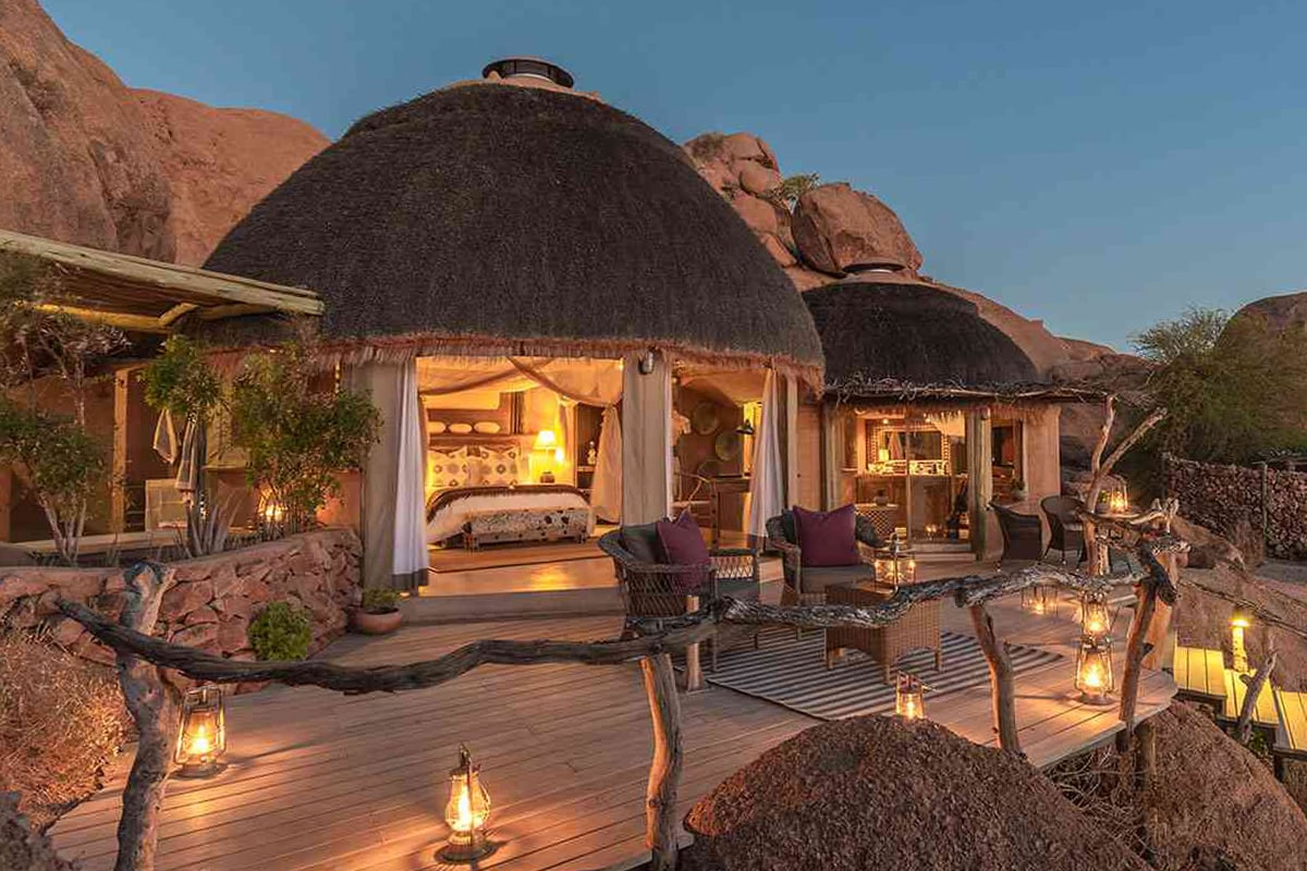 Luxurious Namibia safari lodge at twilight, featuring a thatched roof, open walls adorned with elegant draperies, and stylish outdoor furniture. The warm glow from lanterns illuminates the wooden deck