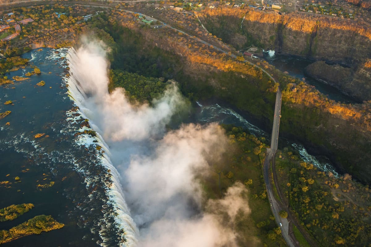 Aerial view of a large waterfall in Zimbabwe with mist rising from its base, surrounded by a green forest and a canyon. A bridge spans the river upstream from the falls.