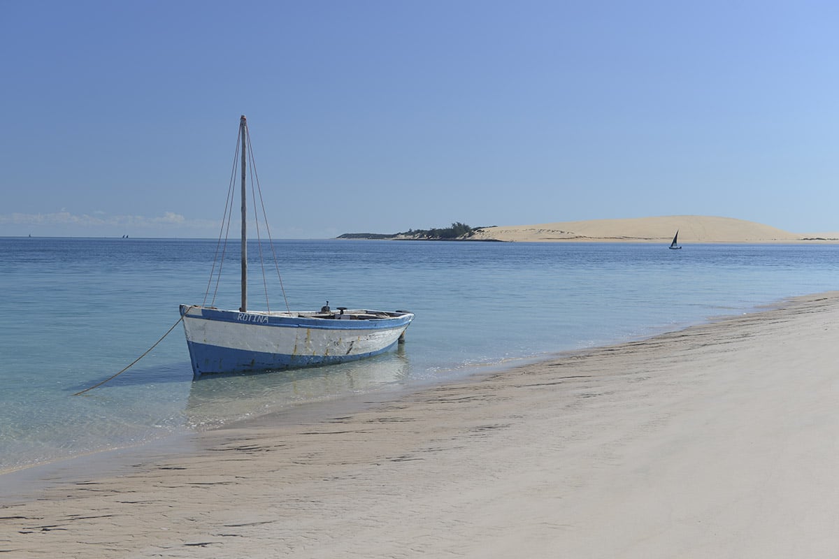 A blue sailboat is moored near the shore of a serene African beach with clear blue waters, overlooking another distant sailboat and a sandy island under a clear sky.
