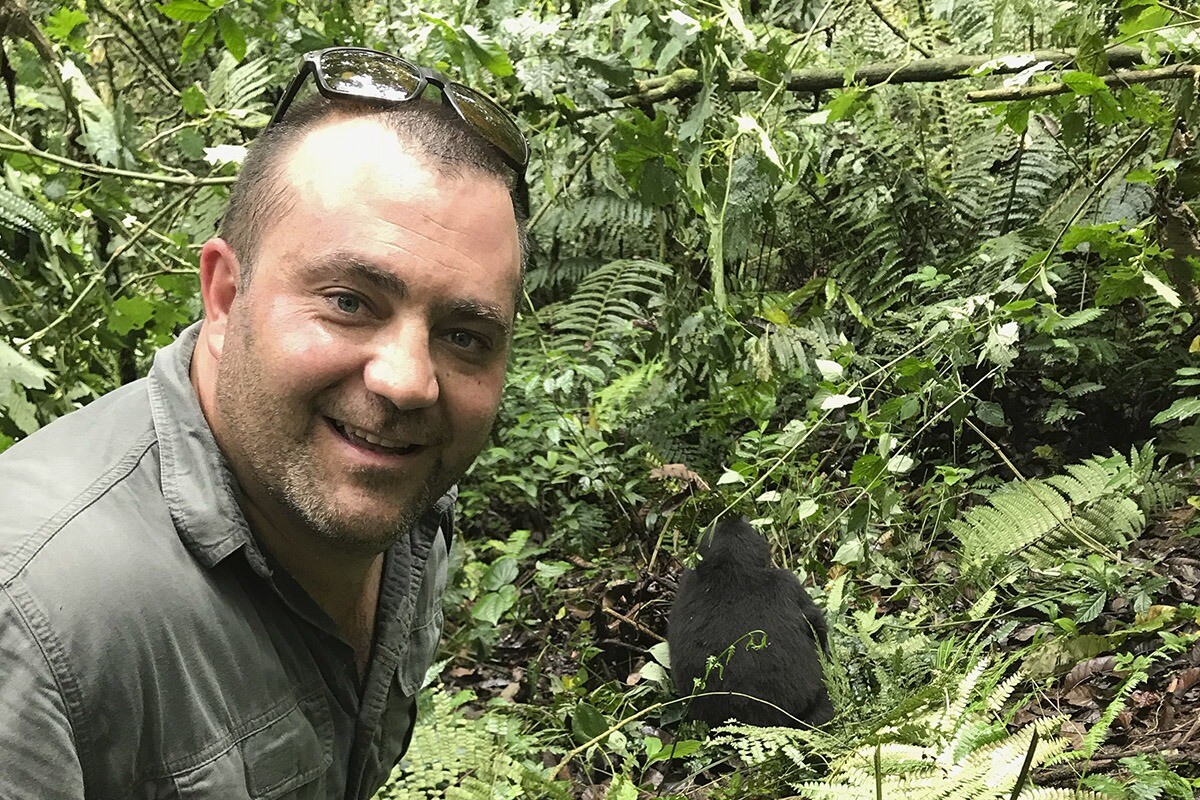 A smiling man with short hair and sunglasses perched on his head posing in a dense jungle environment, with lush green foliage and a small black gorilla visible in the background during a track gorillas tour