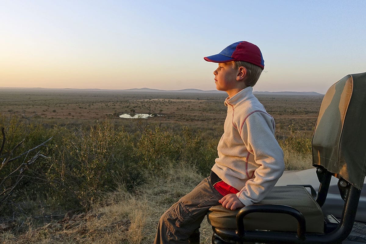 A young boy in a red and blue cap and beige jacket sits on a safari vehicle seat, looking out over a vast, arid landscape with sparse vegetation and distant hills at sunset. A small waterhole is visible in the middle of the scene. The sky is clear with a soft gradient from blue to orange—South Africa, 2019: Travelling With Children.