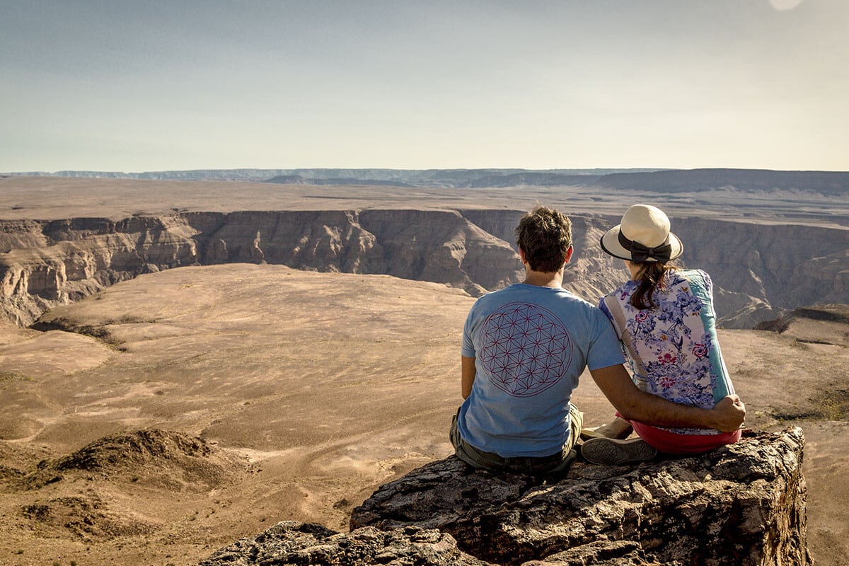 Two people sit on a rocky ledge overlooking a vast, arid canyon landscape in Africa under a clear blue sky. They appear relaxed, one wearing a hat, facing away, enjoying the panoramic view.