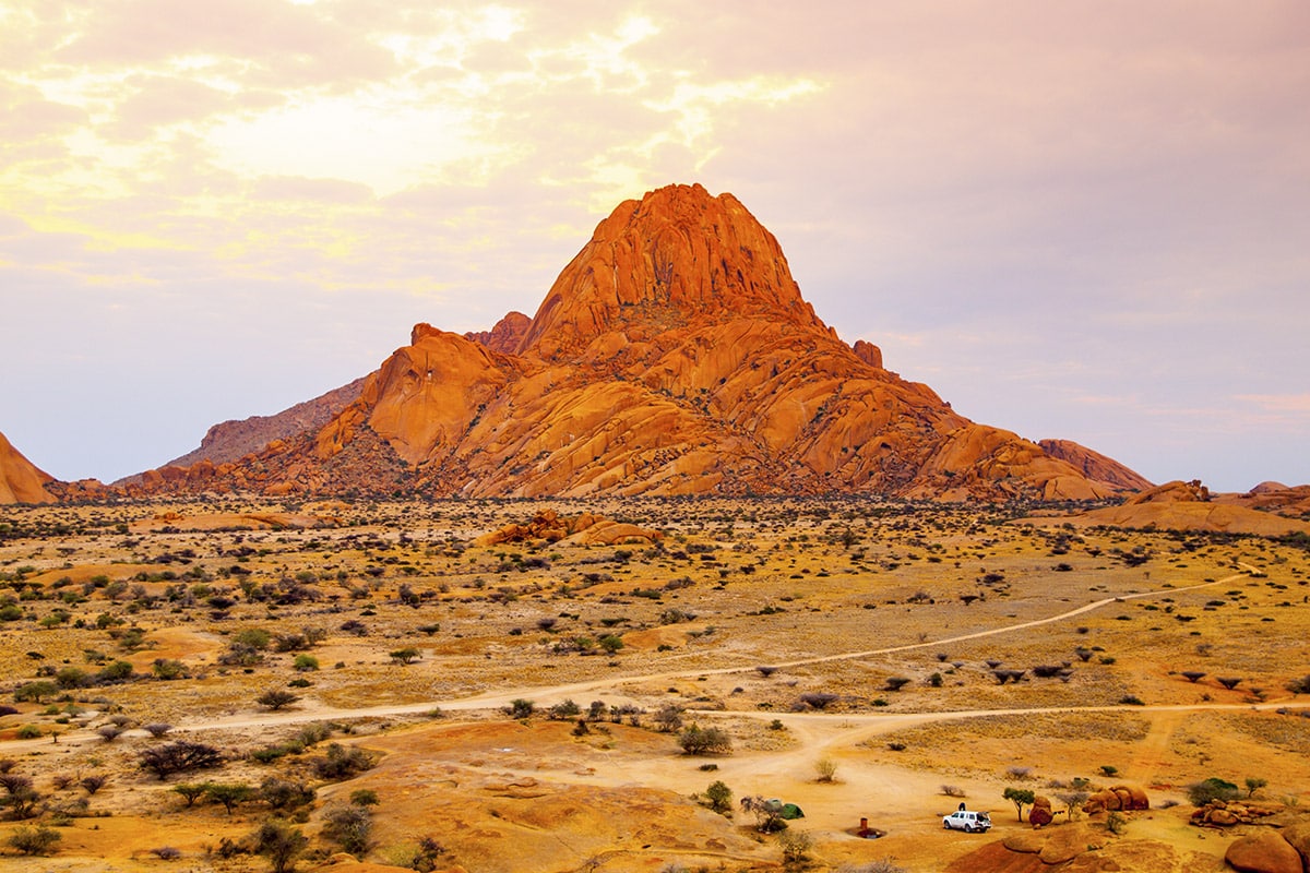 Sunset casts a warm glow on a striking rocky mountain in Namibia's desert landscape, surrounded by sparse vegetation and a clear sky, with a dirt road leading towards it.