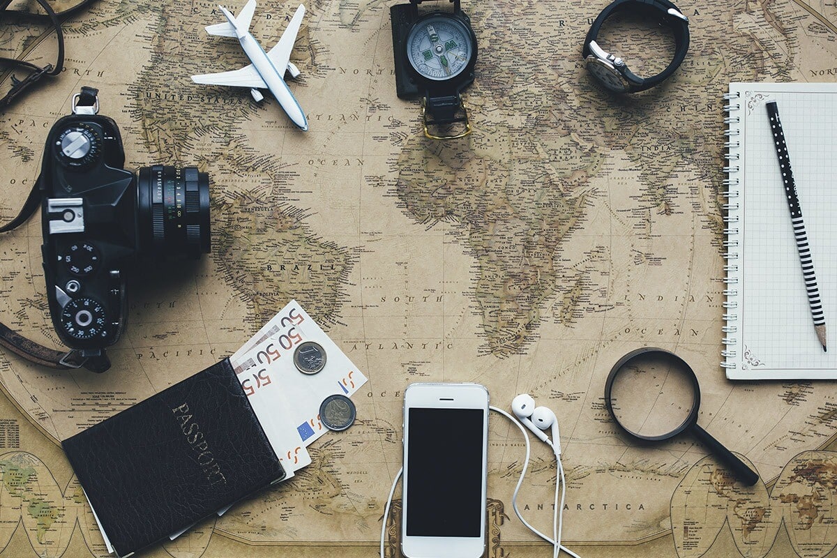 A child-friendly travel-themed layout on a world map includes a camera, compass, magnifying glass, notepad with a pen, passport with foreign currency, airplane model, and smartphone with earphones.
