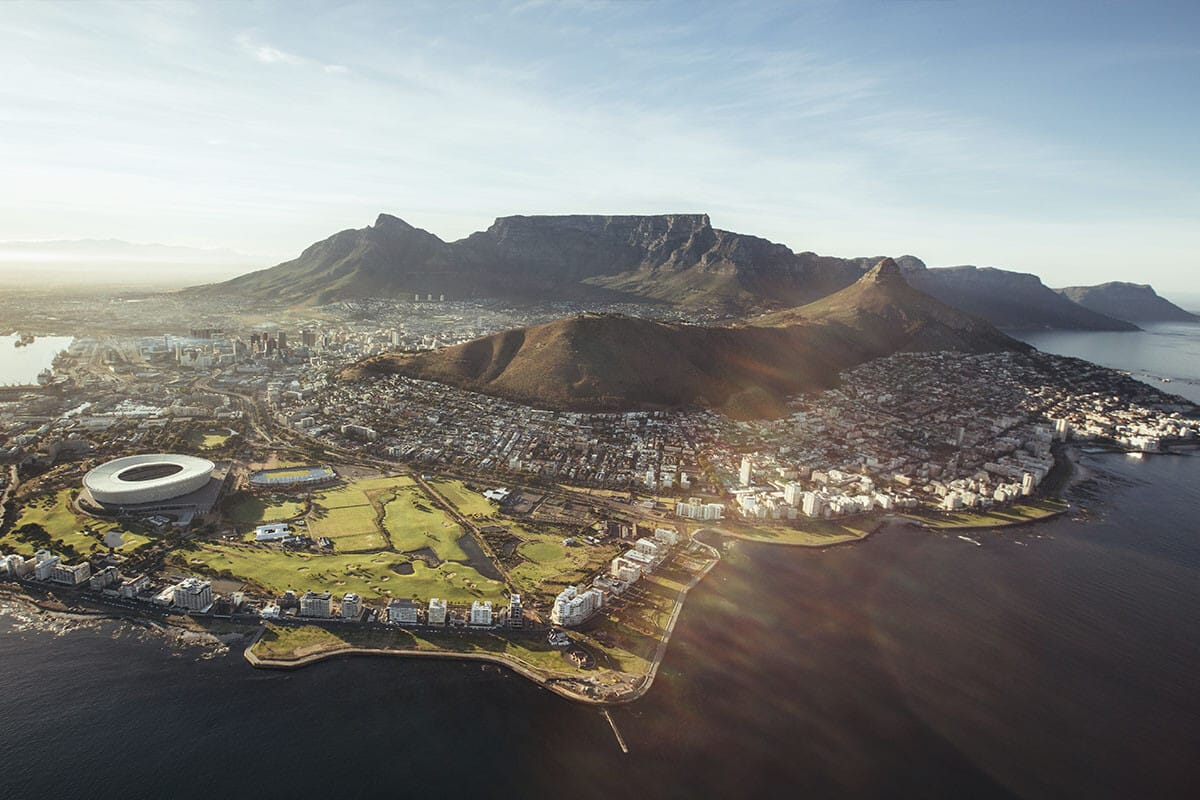 Aerial view of Cape Town, South Africa, featuring Table Mountain in the background, the cityscape, and Cape Town Stadium near the coastline, bathed in the glow of a setting sun with adventure
