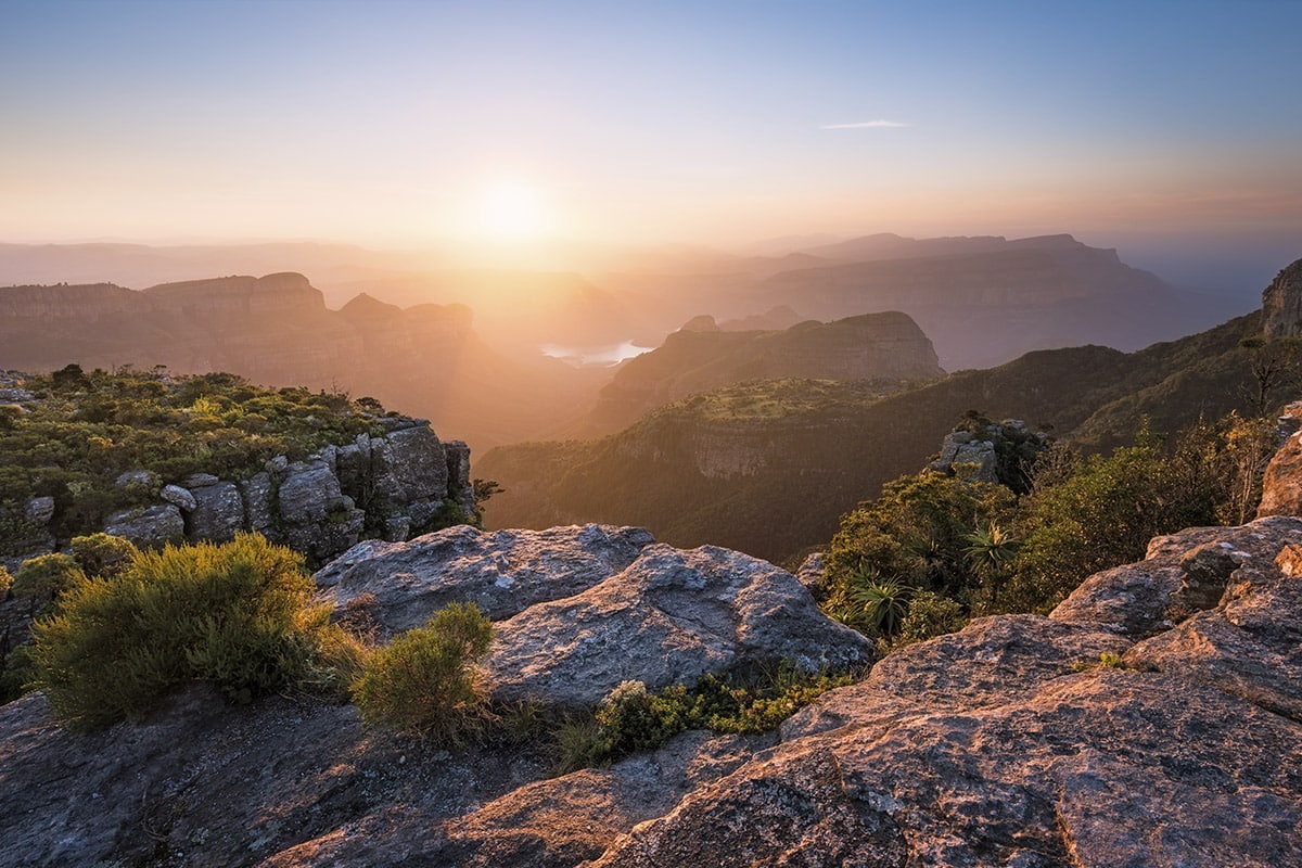 A scenic vista at sunrise showcases a rugged canyon with jagged cliffs and dense vegetation in South Africa. The sun, low in the sky, illuminates the valley and touches the tips of the cliffs,