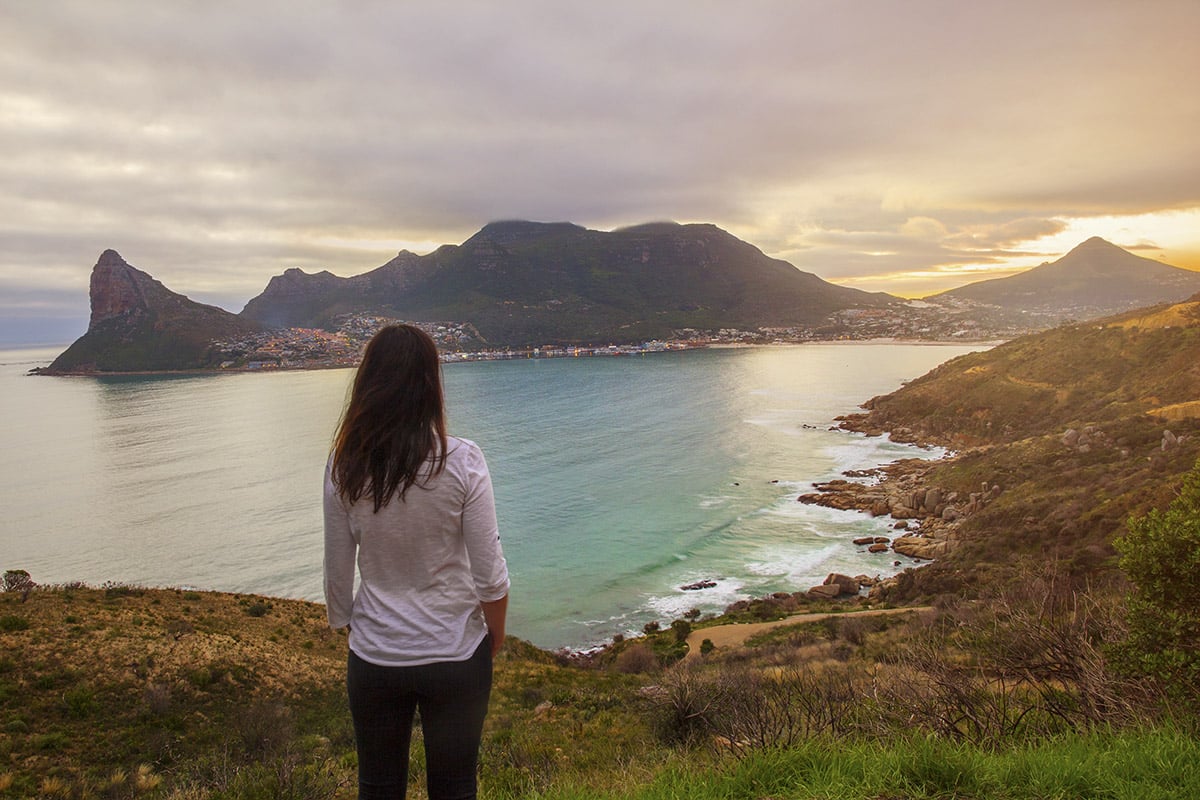 A woman gazes at a scenic coastal view at sunset, featuring undulating mountains and a calm bay. The coastline, ideal for Travelling in Africa, is dotted with lush greenery and rocks under